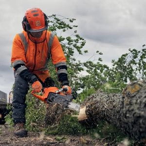 a man dressed in orange chainsaw PPE, using a Husqvarna chainsaw to cut a log on the ground