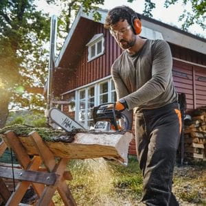 a man using the STIHL MS 162 Petrol Chainsaw to cut firewood on a sawbuck, with a house in the background