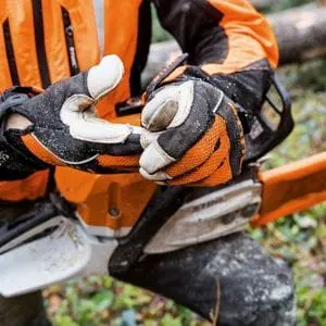 someone dressed in chainsaw PPE, with a chainsaw resting on their knee, taking off a pair of chainsaw gloves