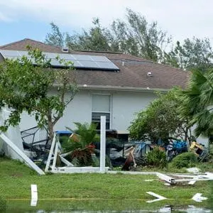 a storm-damaged garden, including fallen trees and debris, in front of a white-walled bungalow with solar panels on the roof