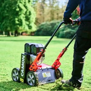 a close up of someone using the Camon ES42 EGO Cordless Scarifier on a lawn, with trees and hedges in the background