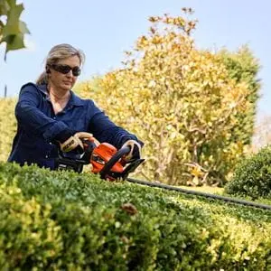 a blonde-haired woman wearing protective sunglasses, using the ECHO DHC-310 Hedge Trimmer to shape the top of a hedge