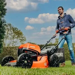 a man with a short beard, using the ECHO DLM 31046SP Lawnmower to mow a lawn, with hedging and trees in the background