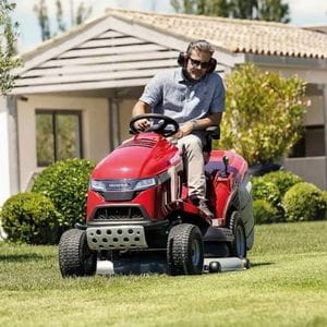 a man wearing ear defenders and protective sunglasses, using the Honda HF 2625 HTE Premium Petrol Lawn Tractor to mow a residential lawn