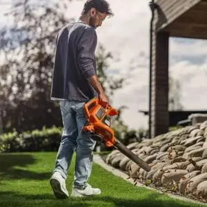 a man using the Husqvarna 120iBV Cordless Handheld Leaf Blower & Vacuum to clear leaves from a lawn, with a decorative stone feature to the right of the picture