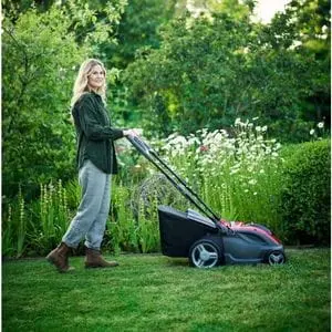 a woman pushing the Mountfield Princess 38 Li Mower across a lawn, with shrubs, hedging and trees in the background