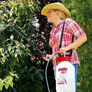 a woman wearing a straw hat and a red checked shirt, using the Solo 461 Comfort Pressure Sprayer to apply treatment to a tall shrub