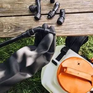 a close up of someone wearing a black glove, holding a sprayer nozzle, with other nozzles on a wooden bench and the sprayer tank on the grass below