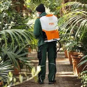 a man wearing a beanie hat, face mask and green overalls, walking between rows of potted plants while using the STIHL SGA 85 Cordless Sprayer to apply treatment to them