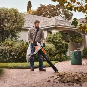 a man using the STIHL SHA 56 Cordless Vacuum Shredder to blow fallen leaves into a pile on a driveway, with a green bag of leaves, a house and trees in the background