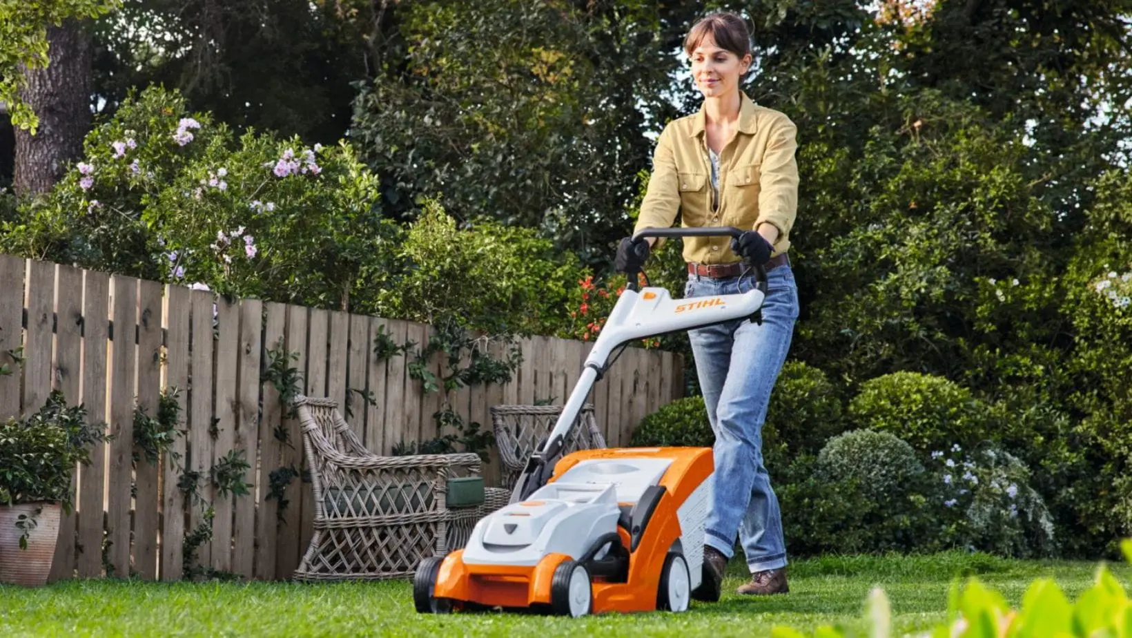 a woman mowing a residential lawn using a STIHL lawnmower, with picket fencing, a wooden garden chair, potted plants, shrubs and trees in the background