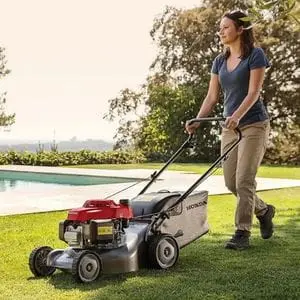 a woman with dark hair, pushing the Honda Izy HRG 416 PK Petrol Lawnmower across a lawn, with a swimming pool and trees in the background