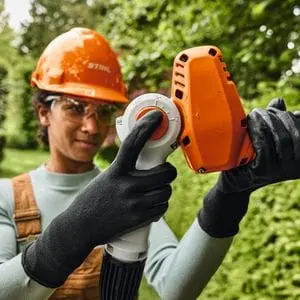 a woman wearing an orange hard hat and protective shades, adjusting the blade on the STIHL HLA 40 Cordless Long Reach Hedge Trimmer 
