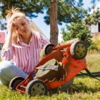 a blonde-haired woman wearing a pink-striped shirt and looking frustrated, sat cross-legged on grass, next to an upturned red lawnmower