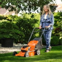 a woman with long hair, using the Husqvarna LC 142i Mower to mow a lawn, underneath an overhanging tree canopy, in a residential area