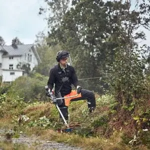 a man wearing a safety helmet and holding a Husqvarna brushcutter, looking at a patch of brambles outside a large white house
