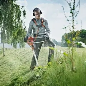 a man wearing safety glasses and a harness, using a Husqvarna brushcutter to clear thick grass and weeds from a slope