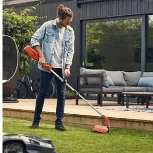 a man operating a Husqvarna strimmer on the edge of a neatly mown domestic lawn, with a modern house and garden furniture in the background and a Husqvarna Automower in the foreground