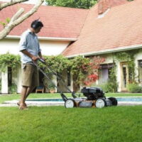 a man wearing headphones and shorts, using a Husqvarna mulching mower to cut a lawn, with a house and swimming pool in the background