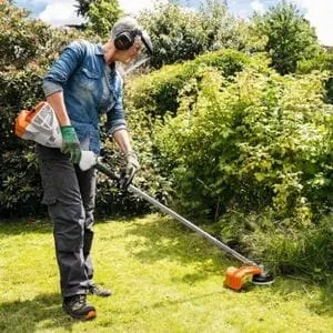a woman wearing a face shield, ear defenders and gloves, using the STIHL FS 56 Petrol Brushcutter to trim tall grass at the edge of a lawn