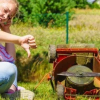 an unhappy looking woman, giving a thumbs-down sign, sat next to an old, upturned electric lawnmower