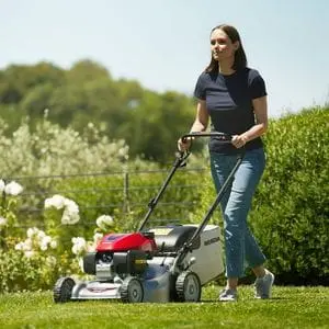 a woman mowing a lawn with a Honda IZY HRG466 Petrol Lawnmower, with plants, hedging and trees in the background