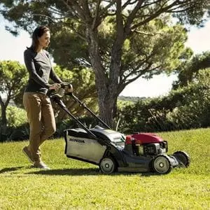 a woman mowing a lawn using the Honda HRX 476 HY Petrol Lawn Mower, with hedging and trees in the background