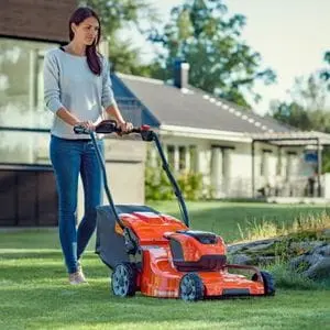 a woman with long dark hair, using a Husqvarna mower to cut a lawn, with houses and trees in the background