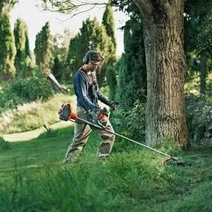 a man using a Husqvarna bike-handle-style brushcutter to cut down rough grass near a tree