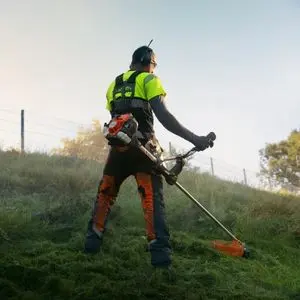 a man facing away from the camera, wearing high-visibility clothing, ear defenders and a strimmer harness, operating a Husqvarna brushcutter on a grassy bank, with a fence at the top of the hill