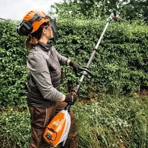 a woman wearing an orange hard hat, using the STIHL HLA 140 Cordless Long-Reach Hedge Trimmer to cut the top of a tall hedge