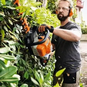 a man wearing protective glasses and ear defenders, using the STIHL HS 82 Petrol Hedge Trimmer to shape the side of a laurel hedge