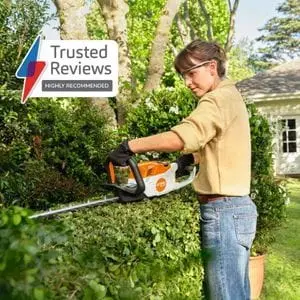 a woman trimming the top of a hedge in a garden using the STIHL HSA 60 Cordless Hedge Trimmer, with a sign in the top left hand corner of the image stating 'Trusted Reviews Highly Recommended'