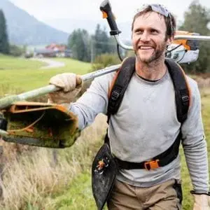 a smiling man, in a picturesque rural scene, carrying a STIHL brushcutter over his shoulder