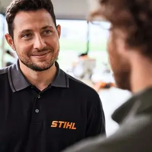 a smiling man wearing a STIHL-branded polo shirt, facing a customer in a garden machinery showroom