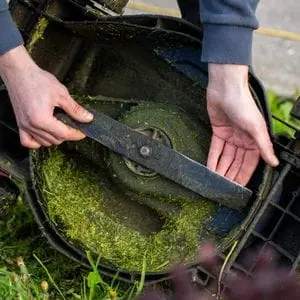 someone holding a lawnmower blade while showing grass buildup on the underside of the deck