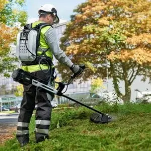 a man wearing a hard hat, high-visibility jacket, work trousers, and an EGO BAX1501 PRO X Backpack Battery, operating an EGO line trimmer on a patch of grass outside a commercial building