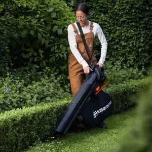 a woman tidying a lawn, near a low-lying hedge, using the Husqvarna Aspire™ BVX-P4A Cordless Handheld Leaf Blower & Vacuum