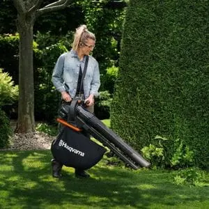 a woman wearing glasses, using the Husqvarna Aspire™ BVX-P4A Cordless Handheld Leaf Blower & Vacuum to clear fallen leaves from a lawn