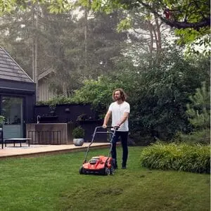 a man with long hair, operating the Husqvarna S 138i Cordless Scarifier on a lawn, with shrubs, trees, a house and patio area in the background