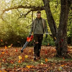 a man walking under a tree, using a Husqvarna leaf blower to clear fallen autumn leaves