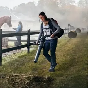 a woman using a Husqvarna blower and vacuum to clear leaves near a horse paddock