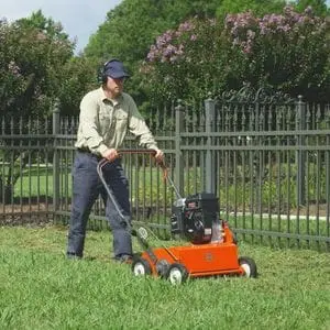 a man operating an orange Husqvarna scarifier on a lawn that is enclosed by a metal, picket-style fence