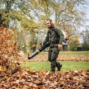 a man wearing ear defenders, using the STIHL BR 800 C-E Petrol Backpack Leaf Blower to move large amounts of fallen leaves in a park