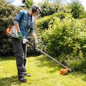 a woman with short grey hair, using the STIHL FS 56 Petrol Brushcutter to trim long grass at the edge of a lawn
