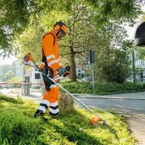 a man wearing orange brushcutter PPE, using the STIHL FS 94 Petrol Brushcutter to cut grass at a roadside