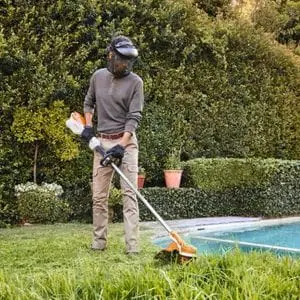a man wearing a faceshield and gloves, using the STIHL FSA 110 Cordless Brushcutter to trim long grass in a garden, next to a swimming pool