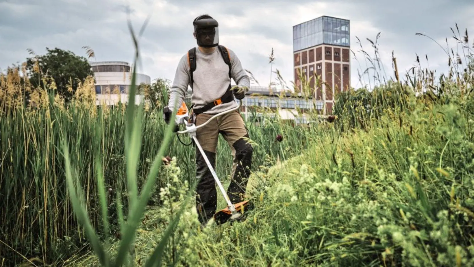 STIHL FSA 140 Cordless Brushcutter (1) a man wearing a faceshield, using the STIHL FSA 140 Cordless Brushcutter to cut vegetation on a slope in front of commercial premises