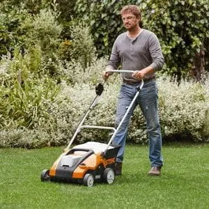 a man pushing the STIHL RLA 240 Cordless Lawn Scarifier across a lawn, with shrubs and trees in the background