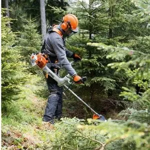 a man wearing a helmet, ear defenders and gloves, using a STIHL brushcutter to cut down vegetation in a clearing in a wood